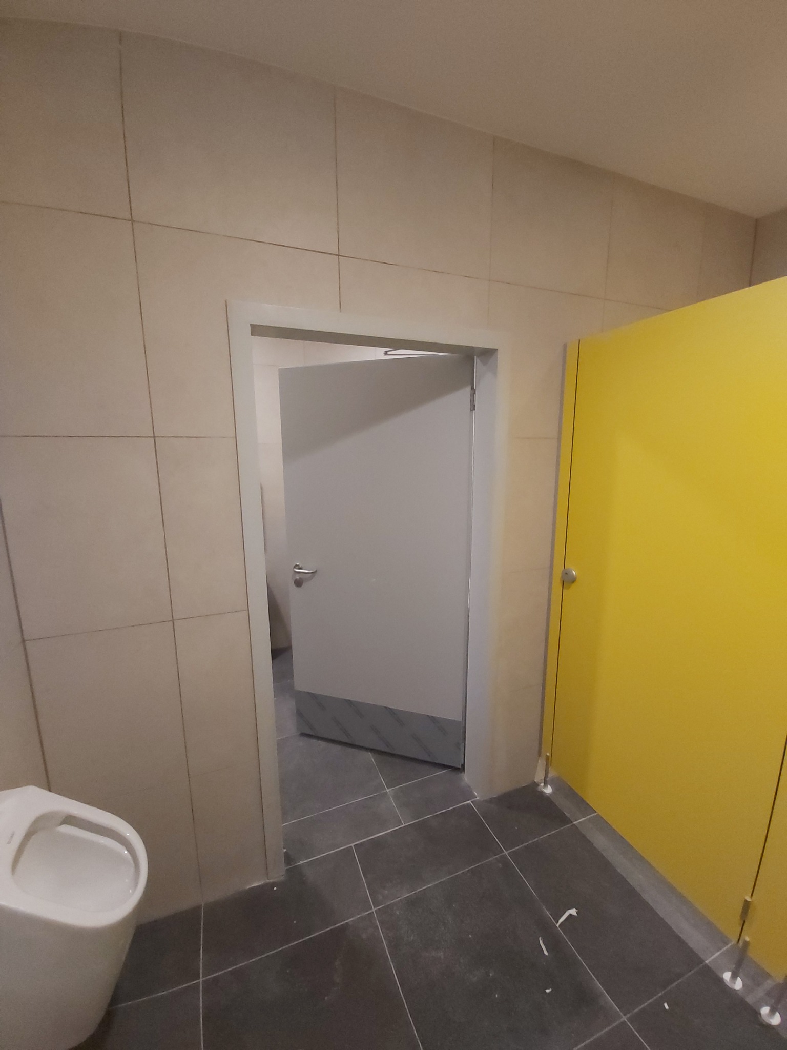 Wnętrze toalety publicznej z beżowymi płytkami ściennymi, szarą podłogą, białą toaletą wiszącą, szarymi drzwiami do kabiny z metalowym wykończeniem oraz żółtą ścianką działową kabiny.