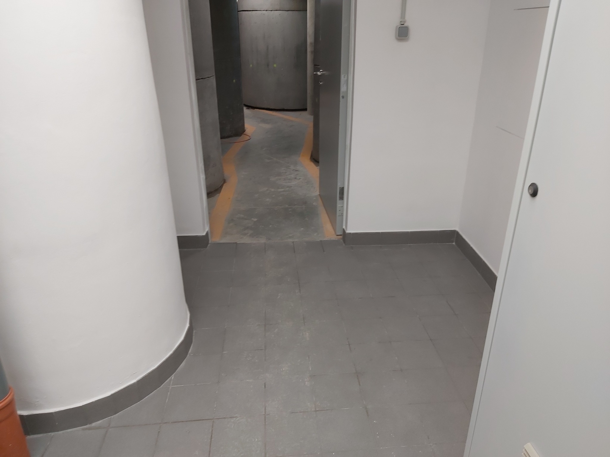 Szare płytki podłogowe w korytarzu z widocznymi betonowymi kolumnami i otwartymi drzwiami.