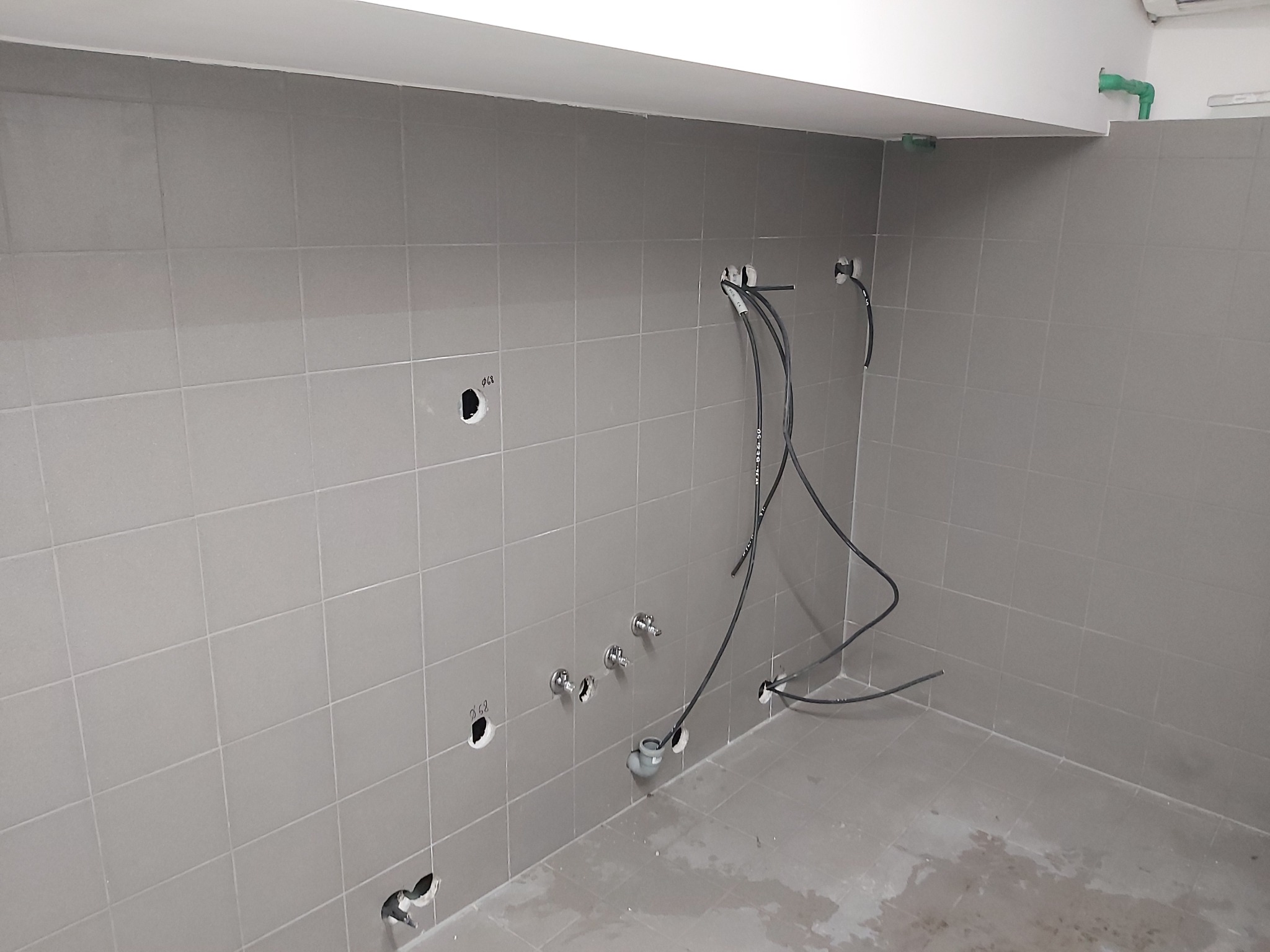 Ściana z szarych płytek w trakcie remontu łazienki lub kuchni, widoczne otwory na instalacje wodne i elektryczne, wystające kable i rury, surowy stan wykończenia.