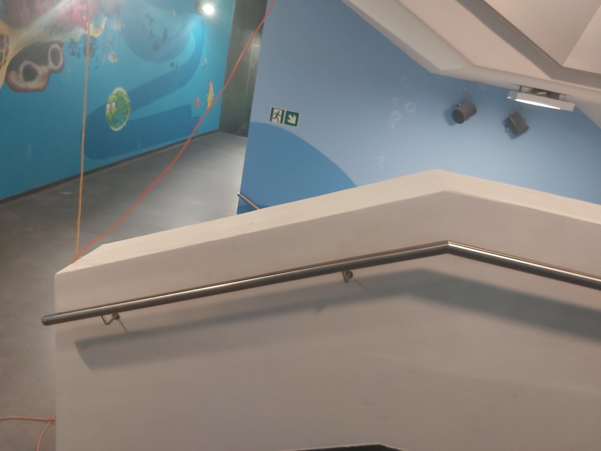 Fragment klatki schodowej z białą ścianą, metalową poręczą i niebieską ścianą ozdobioną motywem podwodnego świata w tle.