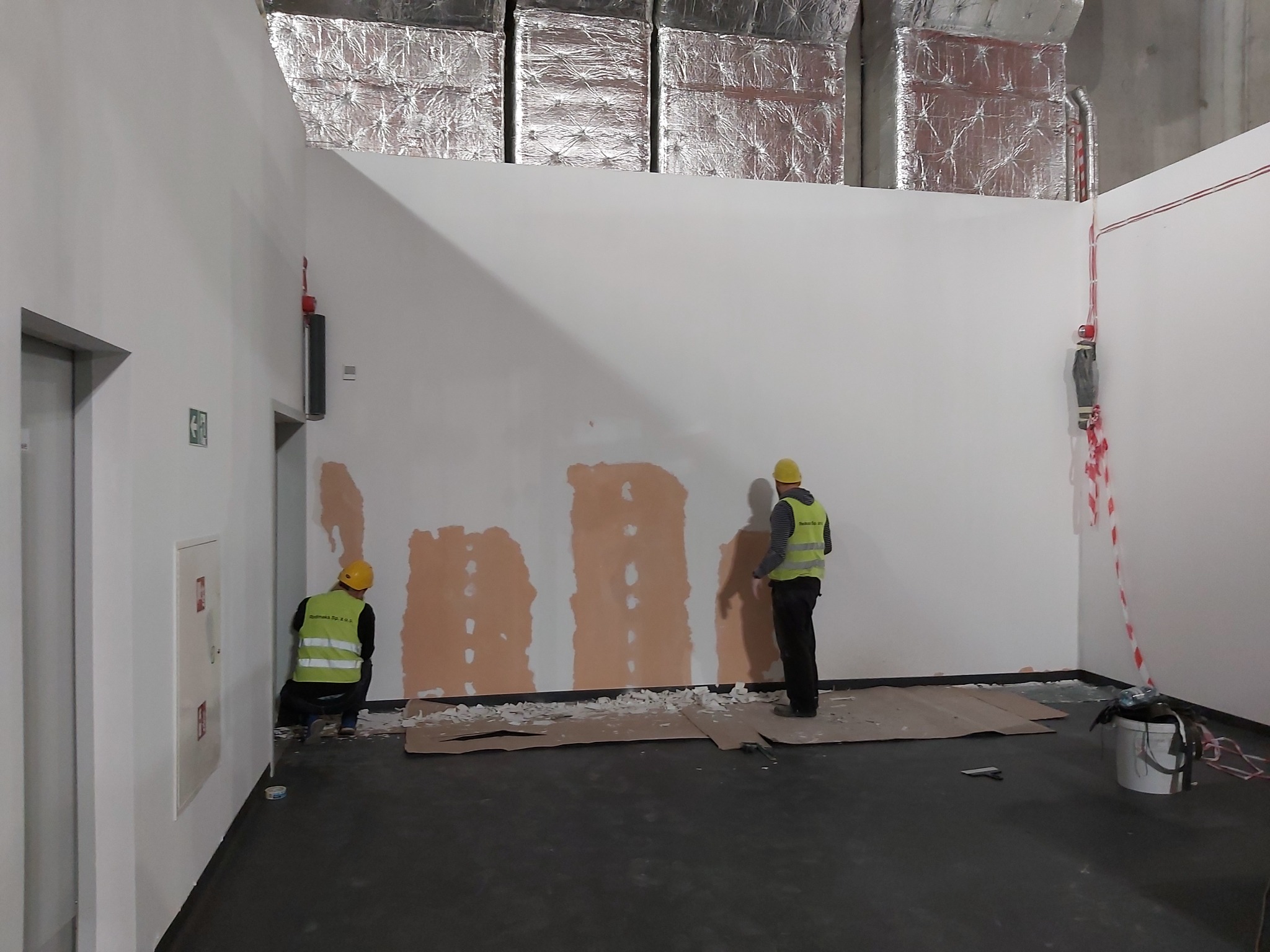 Dwóch pracowników w żółtych kamizelkach i kaskach wyrównuje ścianę w pomieszczeniu z widocznymi instalacjami sufitowymi i zabezpieczeniami przeciwpożarowymi.