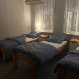 Trzy ł&oacute;żka pojedyncze z drewnianymi ramami i pościelą w odcieniach niebieskiego, ustawione w pokoju ośrodka terapii uzależnień, oświetlone lampkami nocnymi i światłem dziennym wpadającym przez okna...