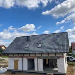 Dom w trakcie budowy z nowym, grafitowym dachem z dachówki ceramicznej, widoczne okna dachowe i komin, budynek z białego bloczka, teren wokół podmokły.