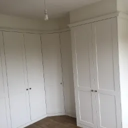 Biała, narożna szafa wnękowa z panelowymi drzwiami i srebrnymi gałkami, dopasowana do kształtu pomieszczenia, z widoczną drewnianą podłogą i żarówką wiszącą na suficie.