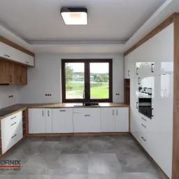 Nowoczesna kuchnia z białymi, lakierowanymi frontami i elementami z drewnopodobnego dekoru, szarymi płytkami na podłodze i oknem z widokiem na zieleń.