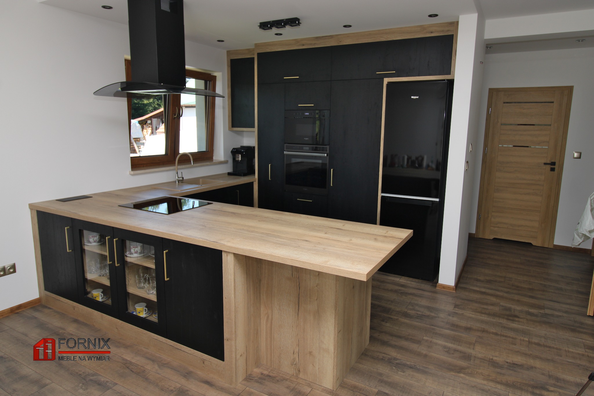 Nowoczesna kuchnia z czarnymi szafkami, drewnianym blatem i wyspą z przeszklonymi szafkami eksponującymi porcelanę, oświetlona światłem dziennym wpadającym przez okno.