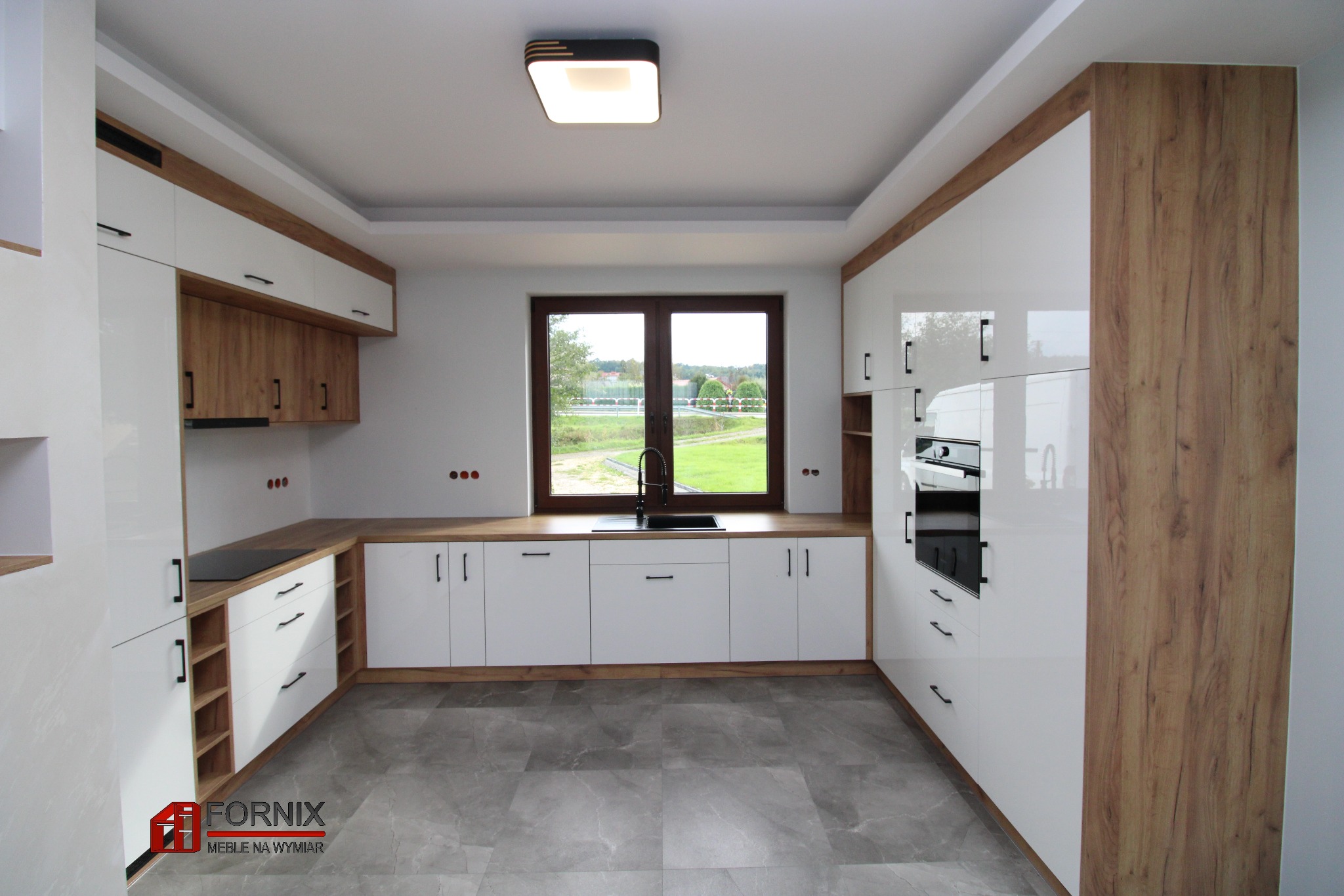 Nowoczesna kuchnia z białymi, lakierowanymi frontami i elementami z drewnopodobnego dekoru, szarymi płytkami na podłodze i oknem z widokiem na zieleń.