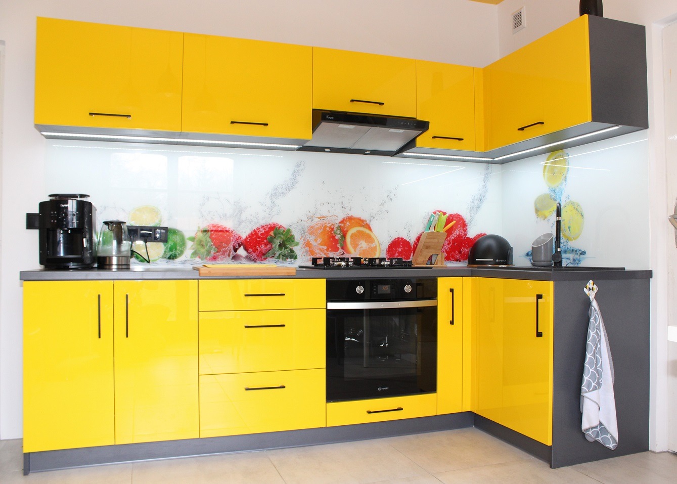 Żółto-szara kuchnia z błyszczącymi frontami, panel szklany z motywem owoców, czarne uchwyty i sprzęty AGD, oświetlenie podszafkowe LED.