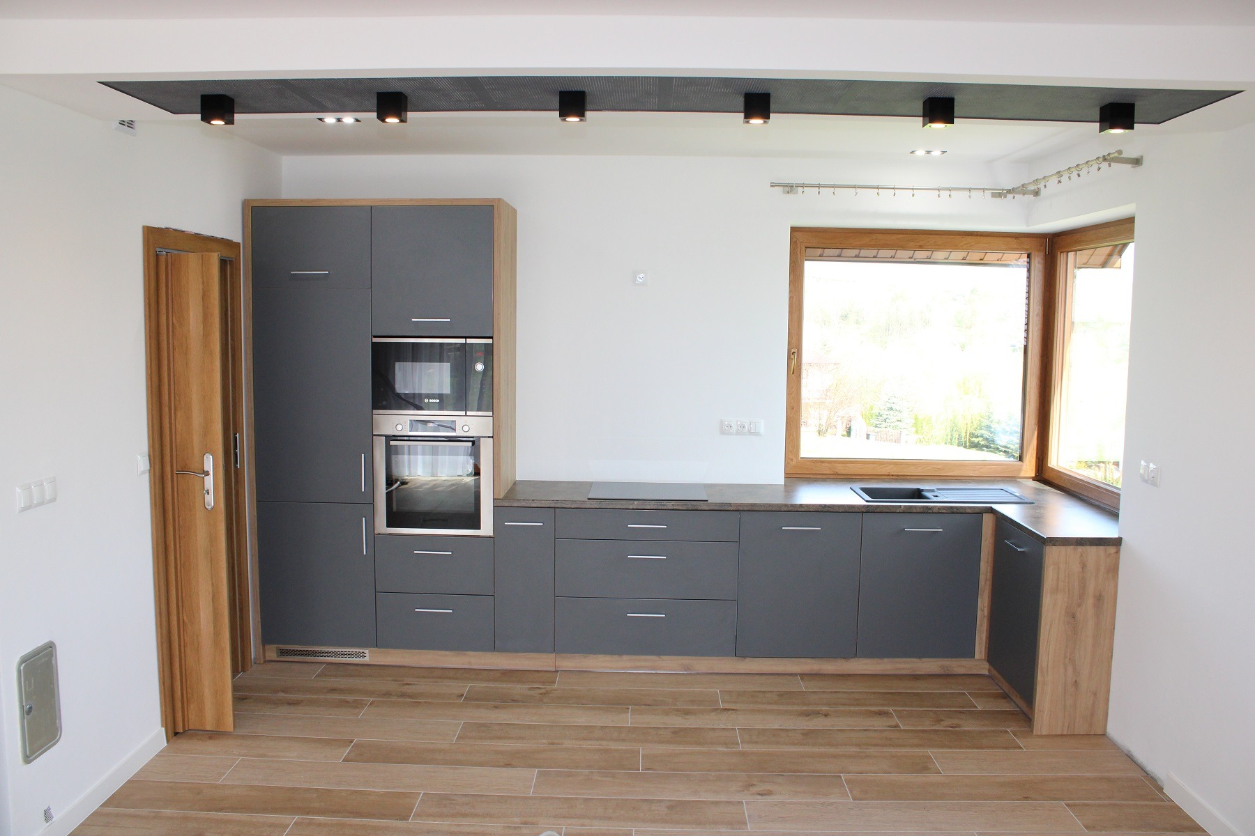 Nowoczesna kuchnia z szarymi frontami szafek, drewnianymi akcentami i oknem narożnym z widokiem na zieleń; oświetlenie punktowe w suficie podwieszanym.