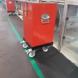 Czerwona metalowa skrzynia transportowa na kółkach z zamkiem szyfrowym w hali produkcyjnej z szarym podłożem i zielonym pasem.