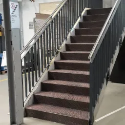 Klatka schodowa w hali produkcyjnej z metalowymi schodami pokrytymi brązową, antypoślizgową wykładziną i szarymi metalowymi balustradami. Na podłodze taśma ostrzegawcza w czarno-żółte pasy.