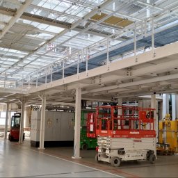 Iron Workers Sp. z o.o. - Wnętrze hali produkcyjnej z metalową konstrukcją nośną pod sufitem, widoczne podesty, instalacje, podnośniki nożycowe oraz elementy wyposażenia.