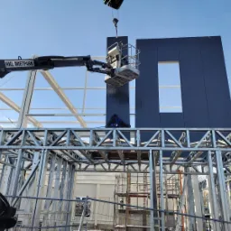 Montaż paneli ściennych z oknem w konstrukcji stalowej hali, widoczny podnośnik koszowy i pracownik zabezpieczony linami, logo HKL Mietpark na ramieniu podnośnika.