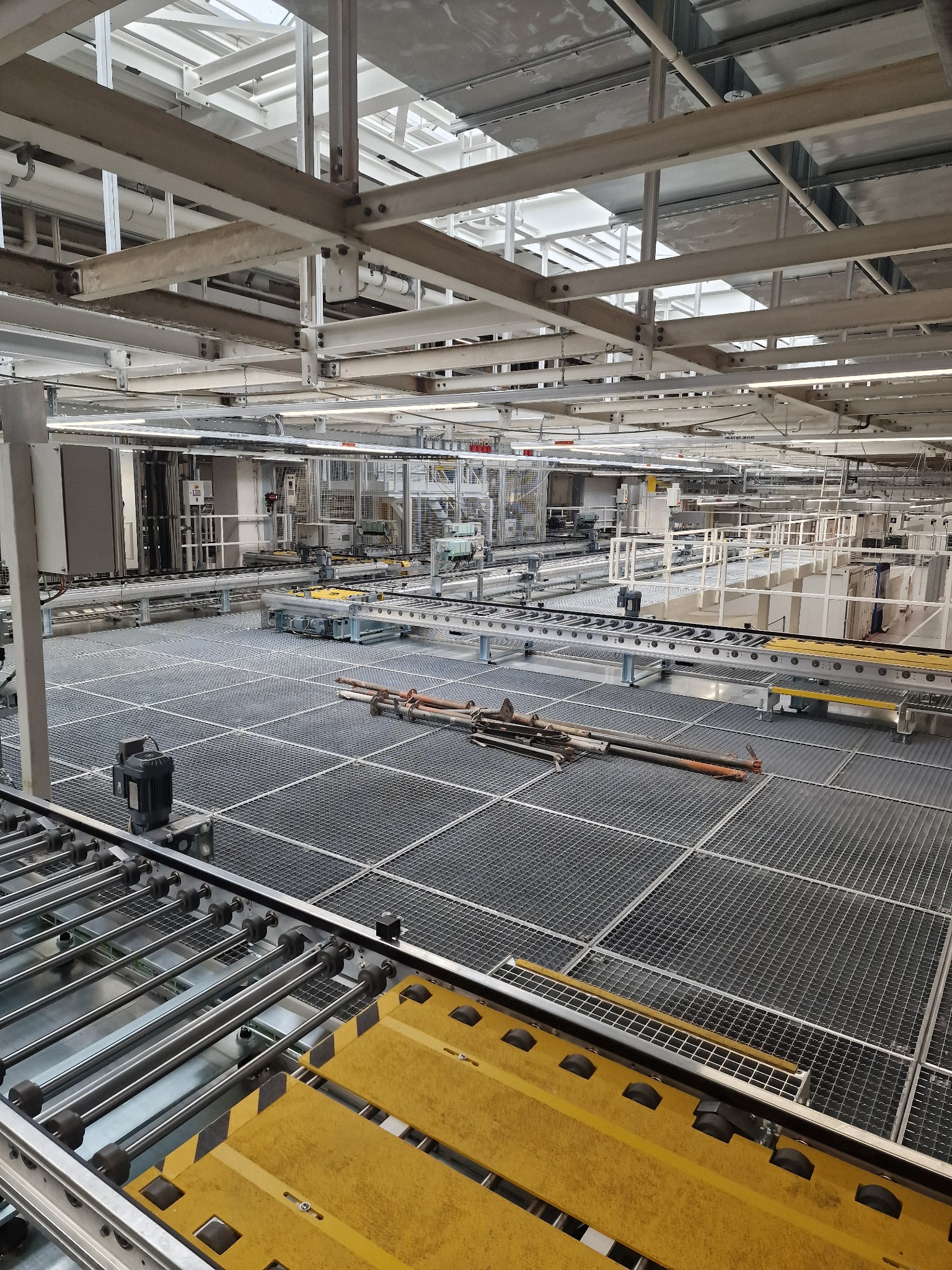 Wnętrze hali produkcyjnej z metalową kratownicą podłogową, widoczne stalowe rury i elementy konstrukcyjne, oświetlenie przemysłowe.