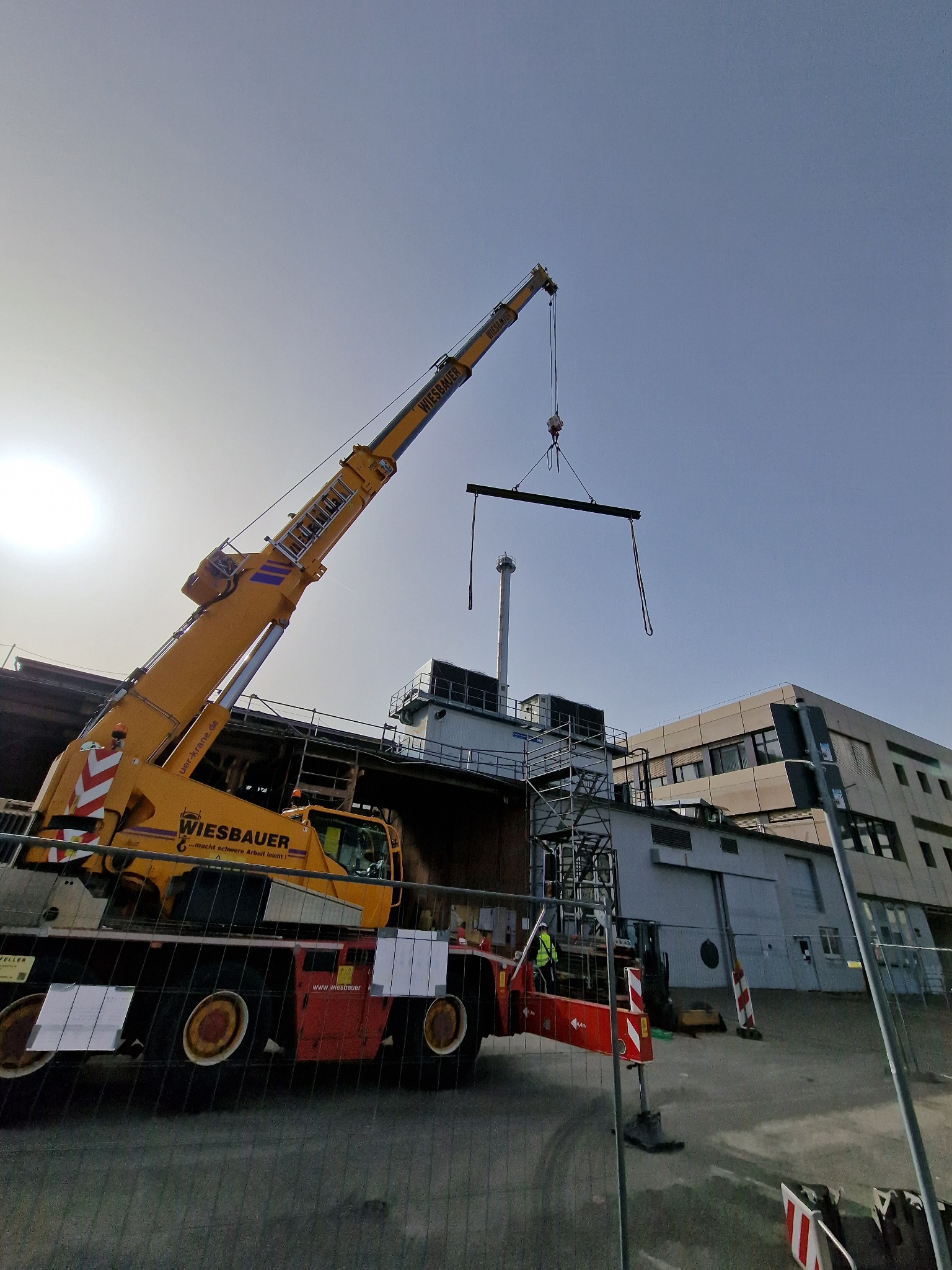 Żółty dźwig Wiesbauer podnosi stalowy element konstrukcyjny na tle budynku przemysłowego i nieba, widoczne rusztowania i pracownik w kamizelce odblaskowej.