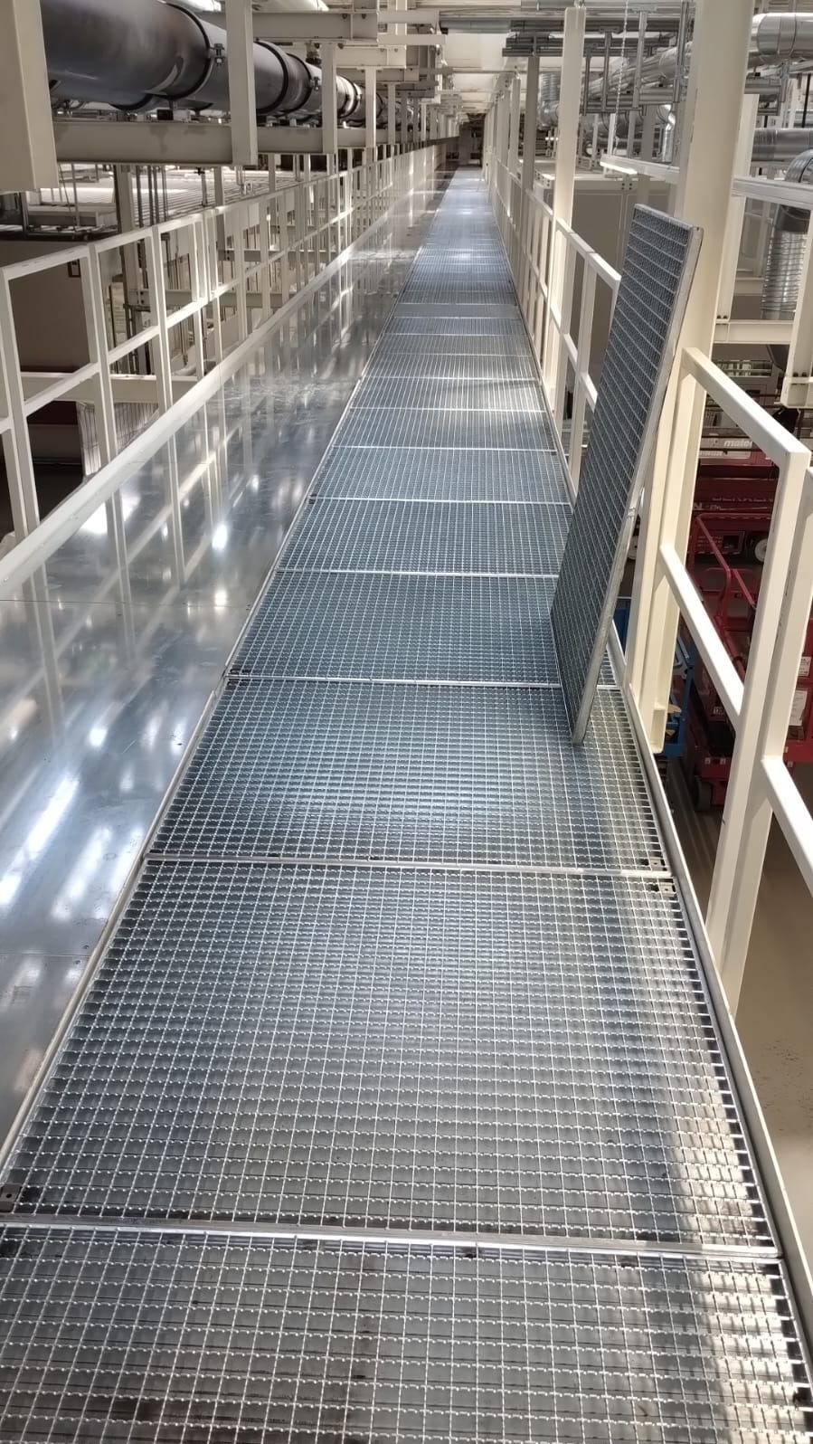 Metalowa kładka z kraty pomostowej, zabezpieczona białą balustradą, w przestrzeni przemysłowej z widocznymi rurami i elementami instalacji pod sufitem.