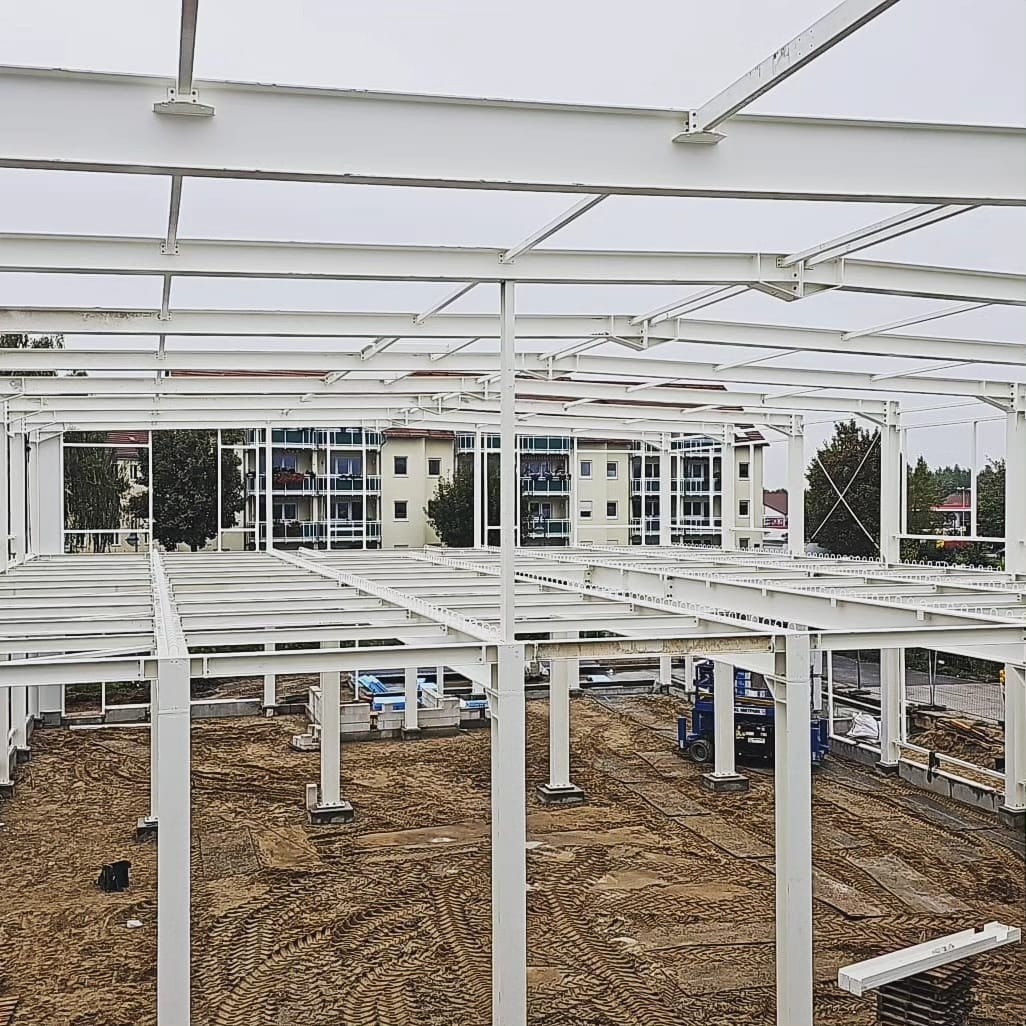 Wznoszenie szkieletu hali stalowej z białych profili, widoczne fundamenty kolumn i niebieski podnośnik koszowy na piaszczystym podłożu, w tle budynek mieszkalny.