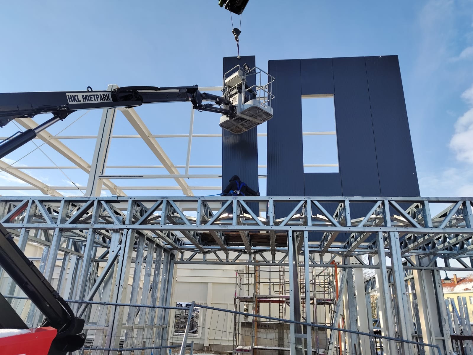 Montaż paneli ściennych z oknem w konstrukcji stalowej hali, widoczny podnośnik koszowy i pracownik zabezpieczony linami, logo HKL Mietpark na ramieniu podnośnika.