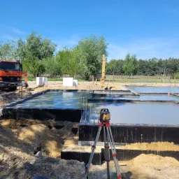 Wylewanie betonu na przygotowane szalunki fundamentowe z widocznym laserowym niwelatorem na statywie i pompą do betonu w tle, w słoneczny dzień.