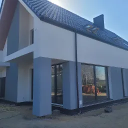 Nowoczesny dom murowany z dwukolorową elewacją (szary i biały), czarną dachówką i dużymi oknami; widoczne przygotowania terenu wokół budynku.