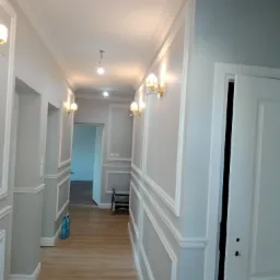 Korytarz po remoncie z panelami podłogowymi, szarymi ścianami z dekoracyjnymi listwami, złotymi kinkietami i otwartymi białymi drzwiami.