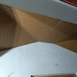 Dom-Bud - Drewniana boazeria na skośnym suficie, ułożona w geometryczny wzór. Ściany pomalowane na biało, widoczne kable elektryczne. Ujęcie z dołu, podkreślające unikalny design.