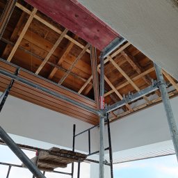 Dom-Bud - Montaż drewnianej podbitki dachowej z rusztowania. Widoczne elementy konstrukcyjne dachu i fragment otynkowanej ściany. Prace wykończeniowe w trakcie realizacji.