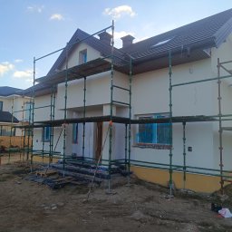 Dom-Bud - Dom jednorodzinny w trakcie prac elewacyjnych, z widocznymi rusztowaniami i zabezpieczonymi oknami. Świeżo otynkowana fasada w jasnym odcieniu, przygotowana do dalszych prac wykończeniowych.