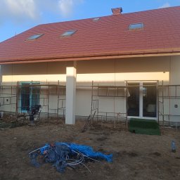 Dom-Bud - Dom jednorodzinny z beżową elewacją i brązowym dachem z lukarnami, w trakcie prac wykończeniowych. Widoczne rusztowania i materiały budowlane.