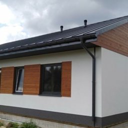 Dom-Bud - Wykończony dom jednorodzinny z drewnianymi okiennicami i elewacją w odcieniach bieli i szarości, widoczny z zewnątrz w pochmurny dzień.