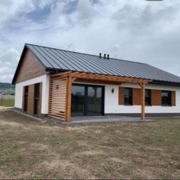 Dom-Bud - Nowoczesny dom jednorodzinny z białą elewacją, drewnianymi elementami dekoracyjnymi i szarą blachodachówką. Widoczna pergola nad tarasem i starannie wykończone detale.