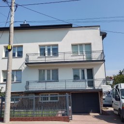 Dom-Bud - Nowoczesny budynek mieszkalny z odświeżoną, gładką elewacją w jasnym kolorze, z dwoma balkonami o kutych, czarnych balustradach. Przed budynkiem stoi biały samochód dostawczy.