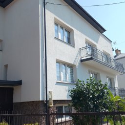 Dom-Bud - Jasny, dwupiętrowy budynek z teksturowaną fasadą, ciemnym dachem i rynną. Na piętrze ozdobny, czarny balkon. Dół elewacji z kamiennym wykończeniem, przed nim metalowe ogrodzenie i zieleń.