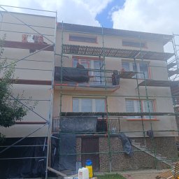 Dom-Bud - Dwupiętrowy dom z rusztowaniem, elewacja w trakcie prac, okna zabezpieczone pomarańczową taśmą. Pracownik na balkonie, kamienna podmurówka, widoczne narzędzia.