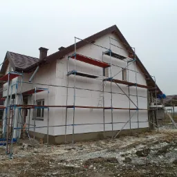 Dom jednorodzinny w trakcie prac z użyciem rusztowania, ściany pokryte białymi płytami izolacyjnymi, widoczni pracownicy w odzieży roboczej, teren wokół budynku częściowo zabłocony.