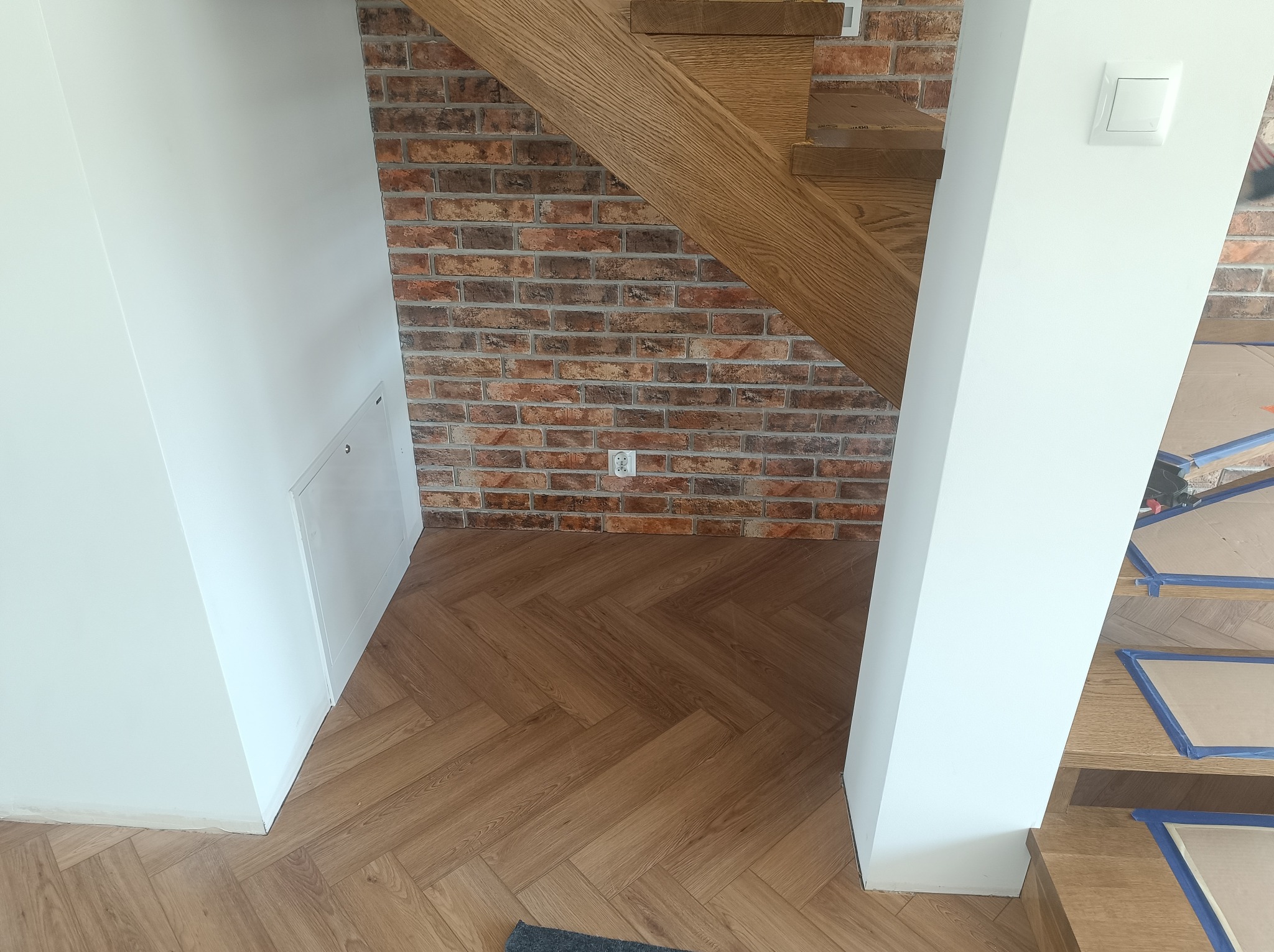 Wnętrze domu z drewnianymi schodami, parkietem ułożonym w jodełkę i fragmentem ściany z cegły dekoracyjnej, białe ściany i drzwi do schowka.