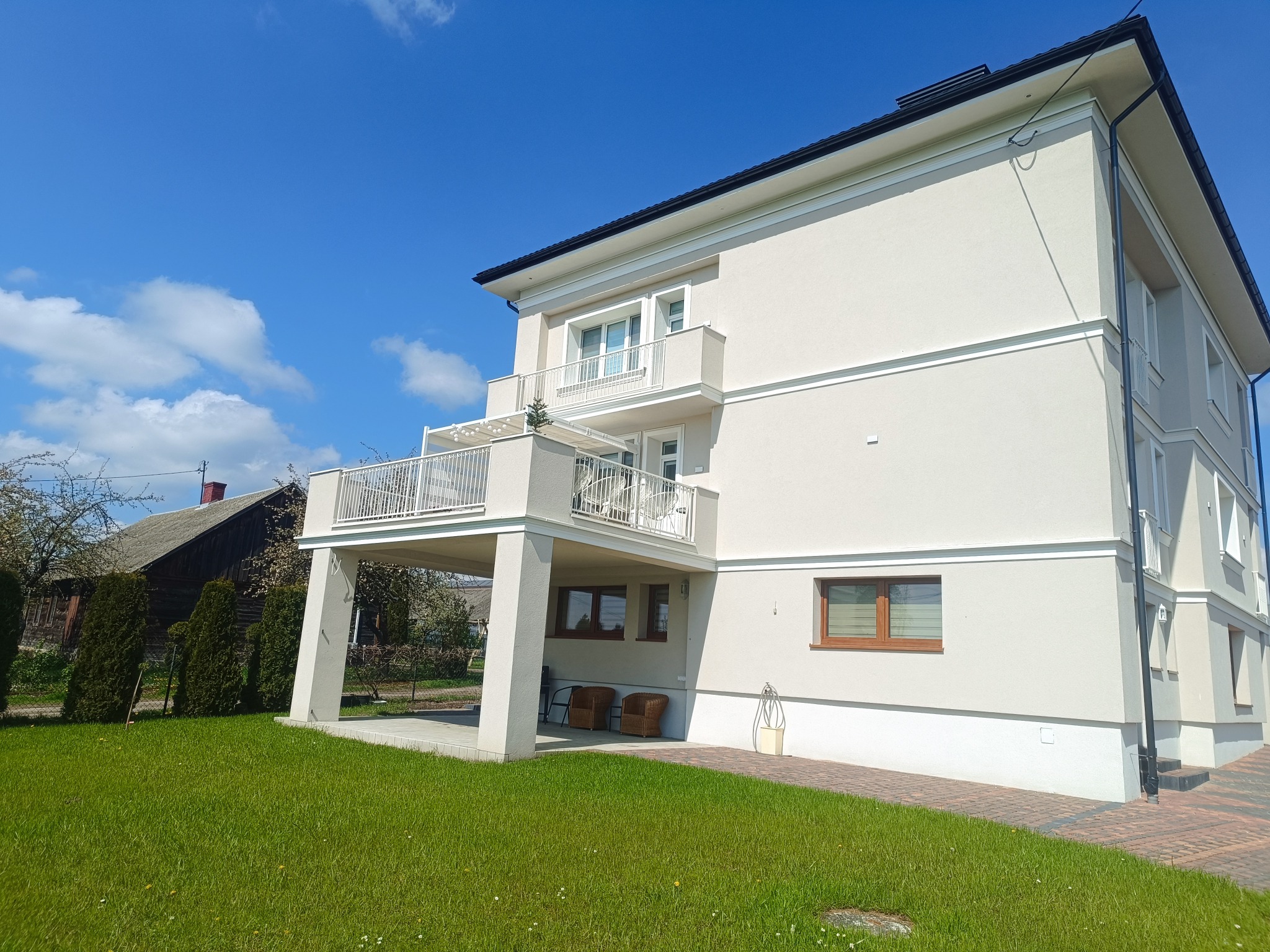 Jasny, dwupiętrowy dom z białymi balkonami i elewacją w odcieniach beżu, otoczony zielonym trawnikiem i starą, drewnianą zabudową w tle pod błękitnym niebem.