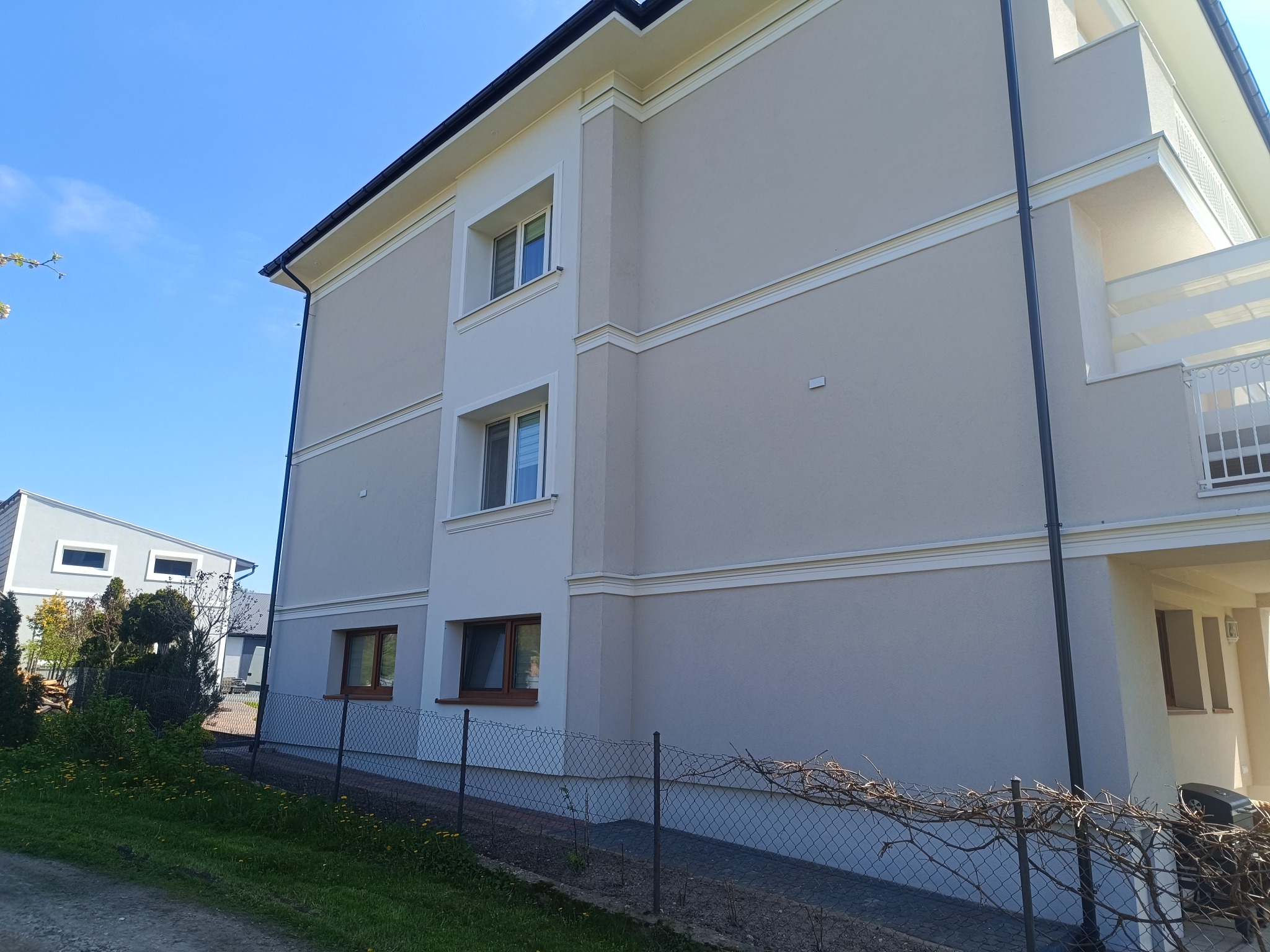 Jasna elewacja dwupiętrowego budynku z białymi detalami architektonicznymi, widoczna zza metalowego ogrodzenia i zieleni.