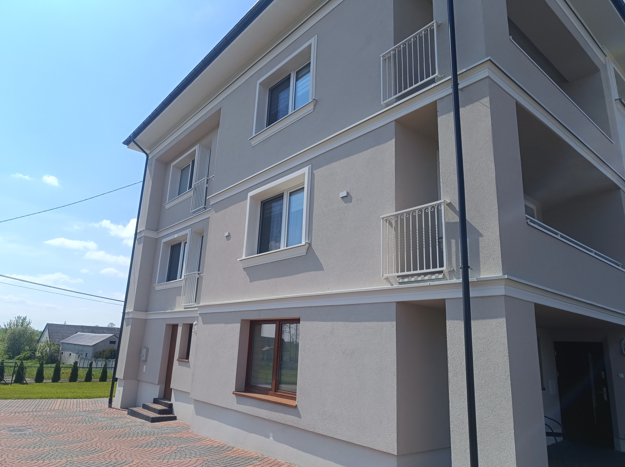 Wykończona elewacja budynku wielorodzinnego z balkonami, w jasnych odcieniach szarości, z białymi detalami architektonicznymi i brązowymi ramami okiennymi, widoczna z perspektywy chodnika...