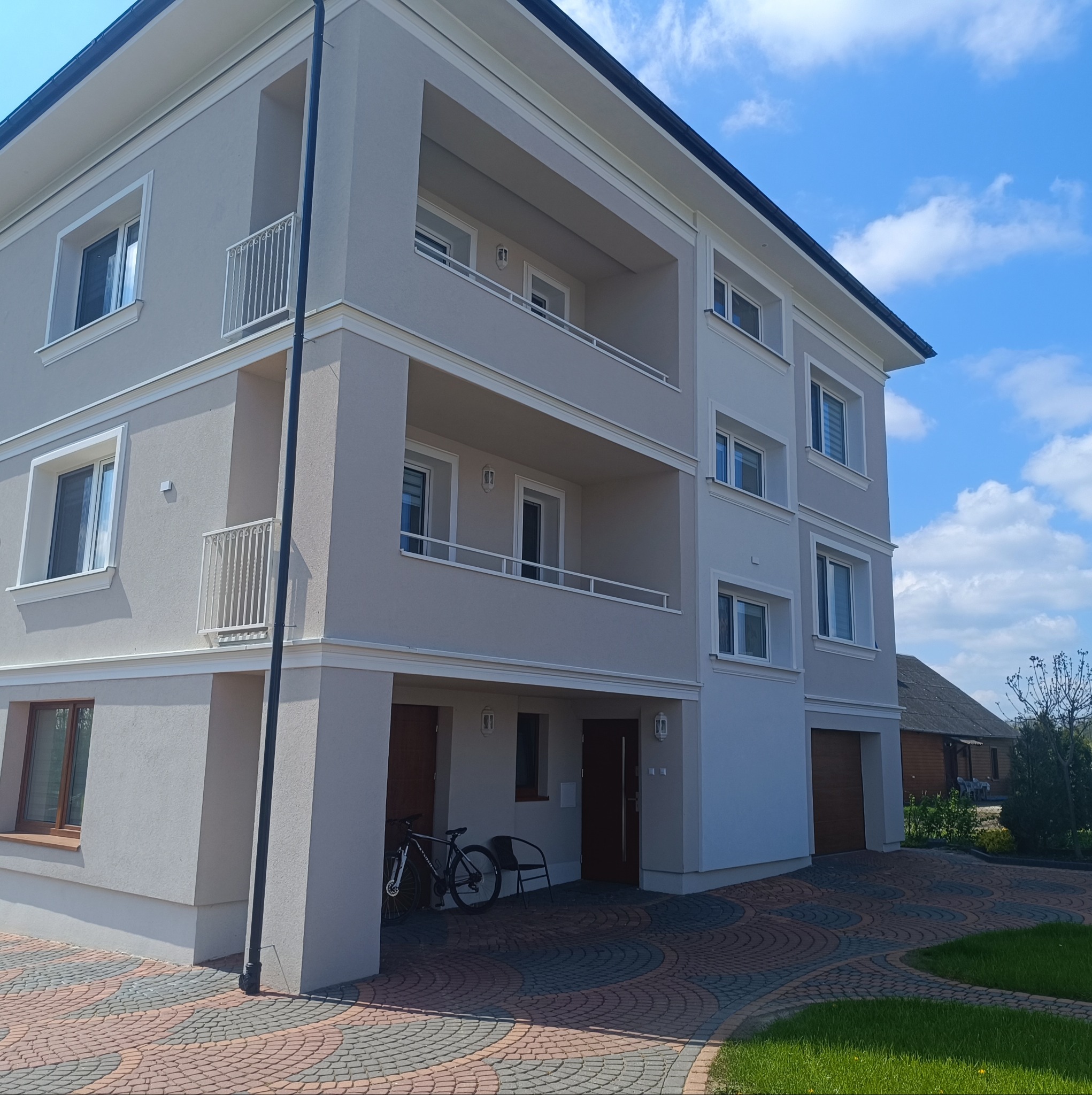 Nowoczesny, trzypiętrowy dom z balkonami w Kleosinie, z elewacją w odcieniach szarości i beżu, sfotografowany w słoneczny dzień z perspektywy chodnika wyłożonego kostką brukową.