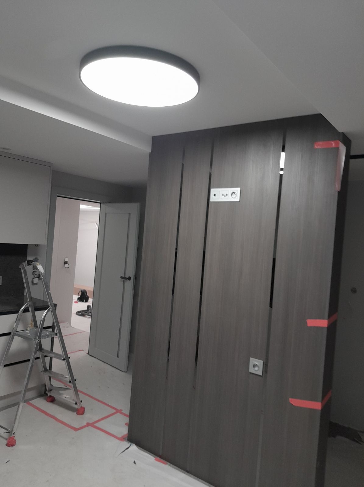 Montaż ścianki działowej z płyty karton gips z widocznymi pionowymi szczelinami dekoracyjnymi, zabezpieczona taśmą malarską, na podłodze widoczne ślady po wyznaczaniu położenia, drabina w tle.