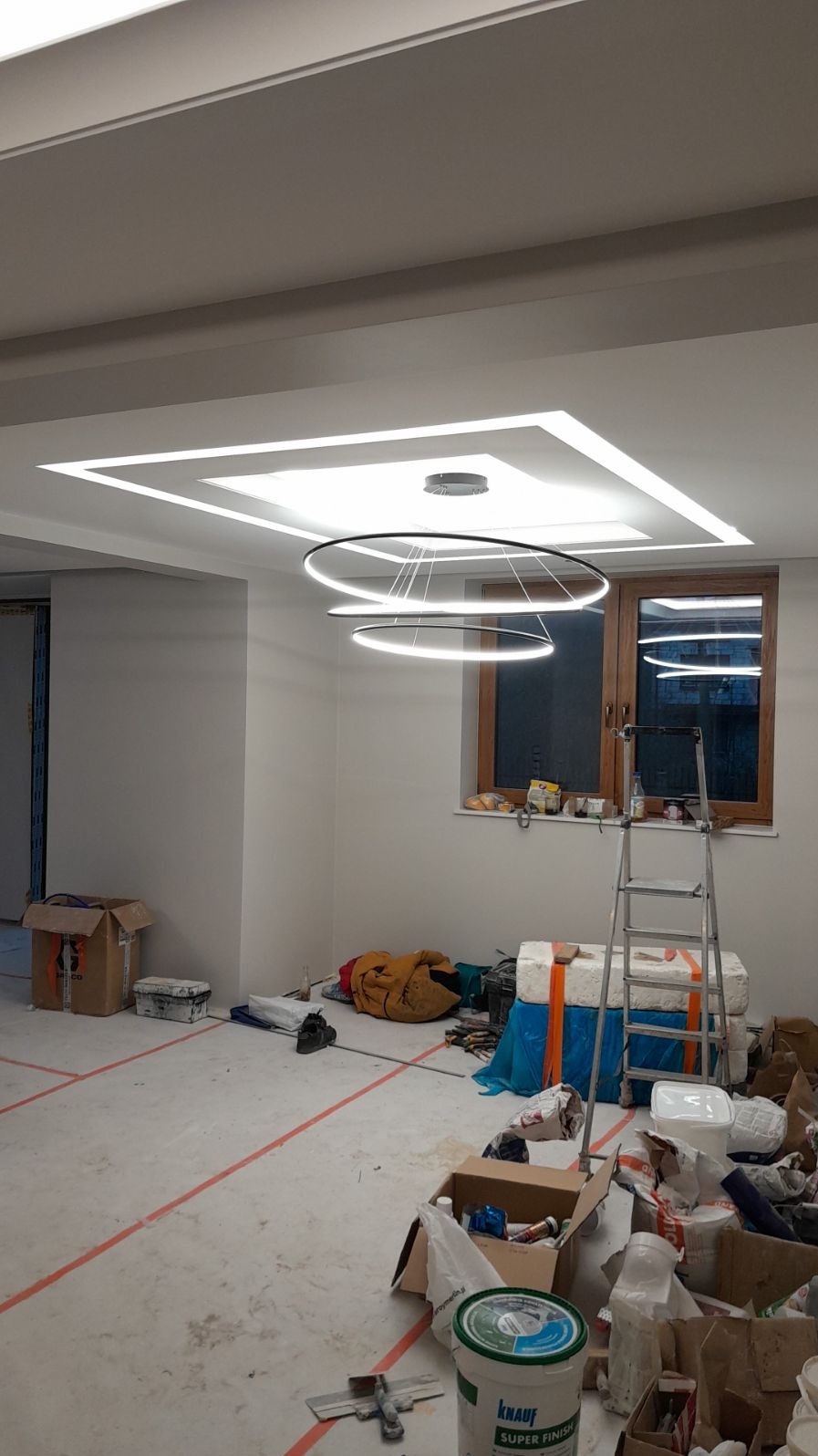 Wnętrze w trakcie wykańczania, widoczne gładkie ściany, sufit z oświetleniem LED w geometrycznym wzorze, podłoga z widocznymi śladami prac, drabina oparta o okno i materiały budowlane. Na pierwszym...