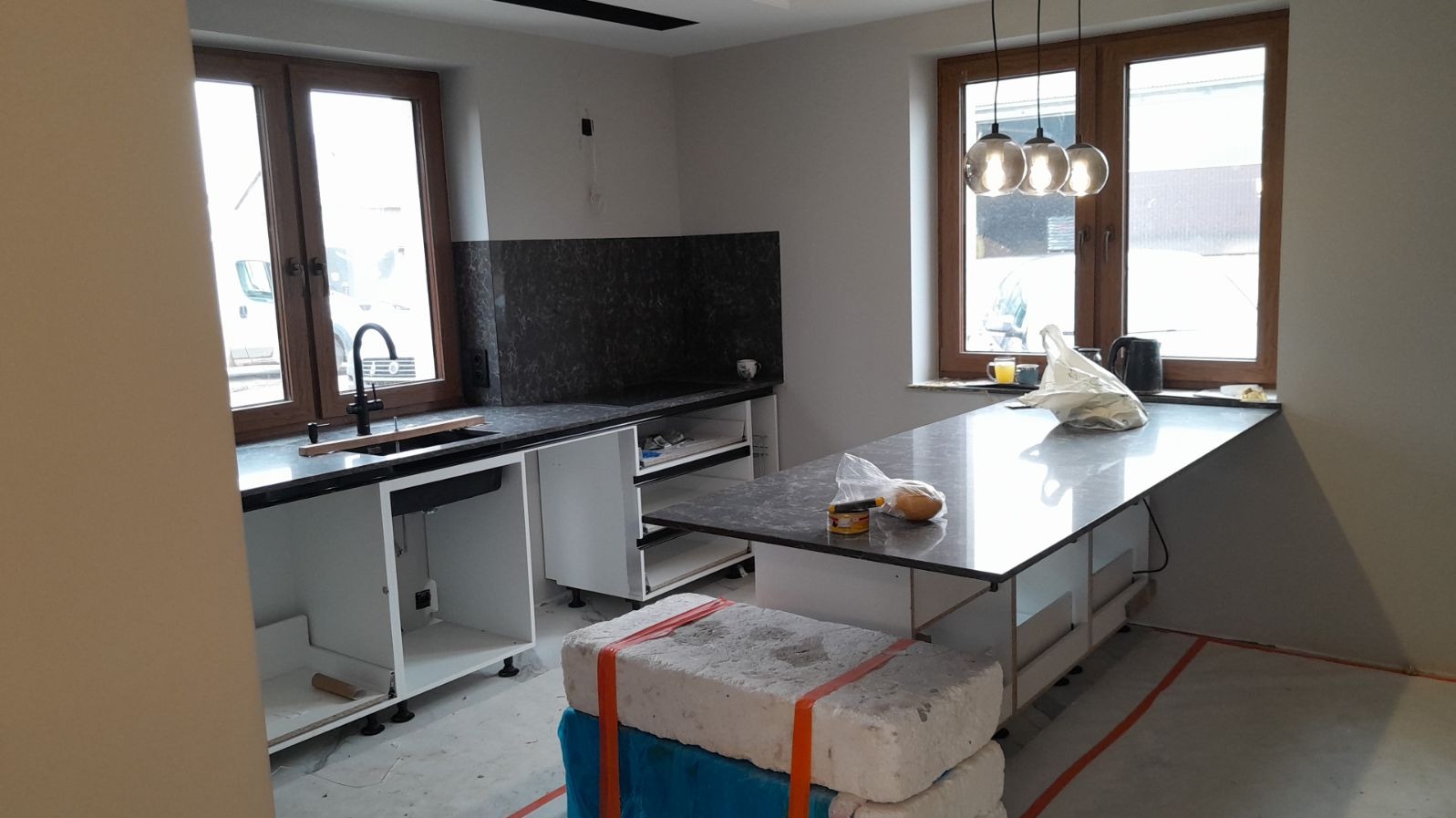Wnętrze kuchni w trakcie montażu: widoczne niedokończone szafki z czarnym blatem, granitowy panel na ścianie, okna z drewnianymi ramami, trzy wiszące lampy i materiały budowlane na podłodze.