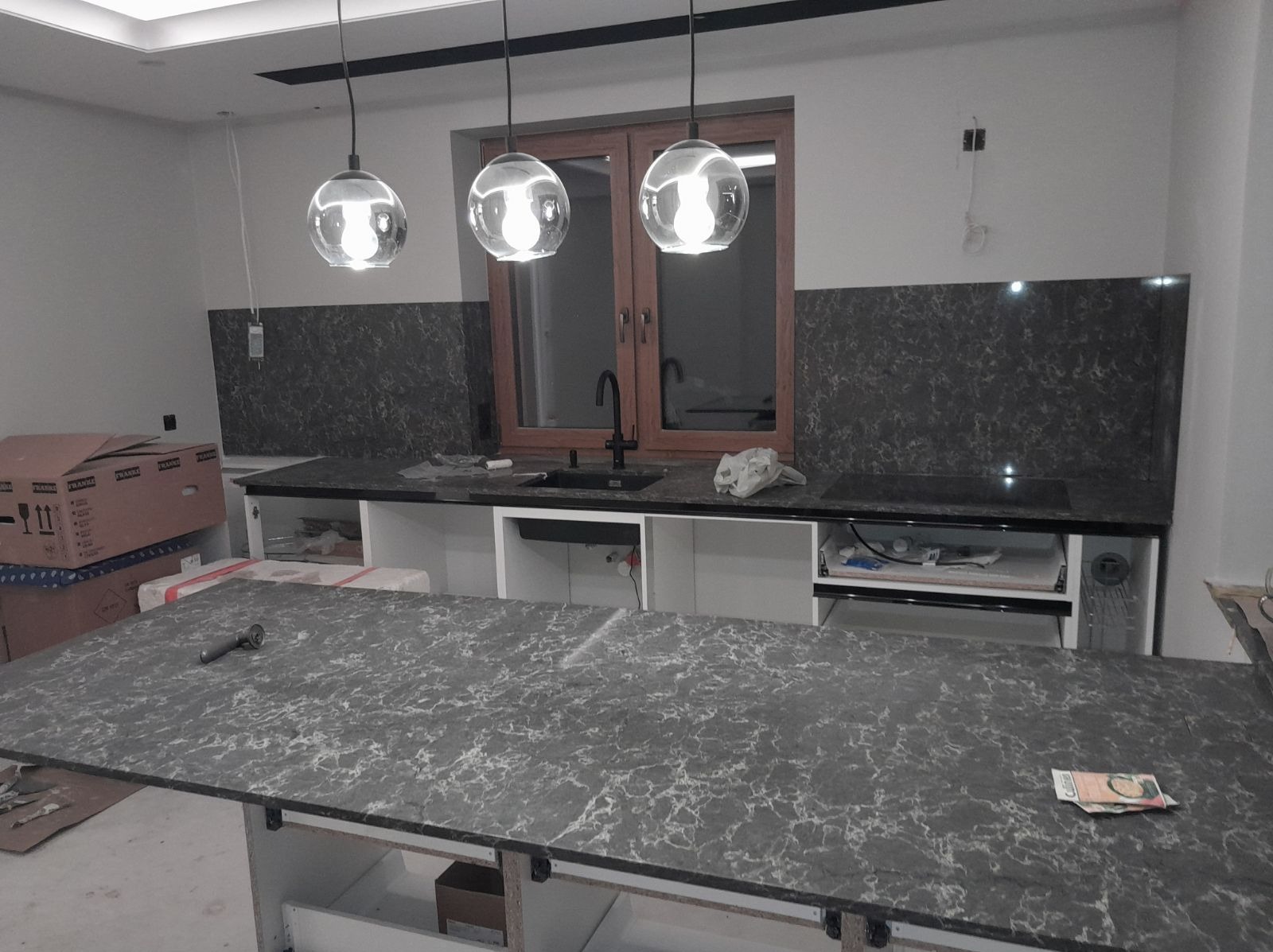 Wnętrze kuchni w trakcie montażu, widoczny blat z szarego kamienia z białymi żyłkami, trzy szklane lampy wiszące, okno w drewnianej ramie i fragment ściany z szarymi płytkami. Na blacie leżą...