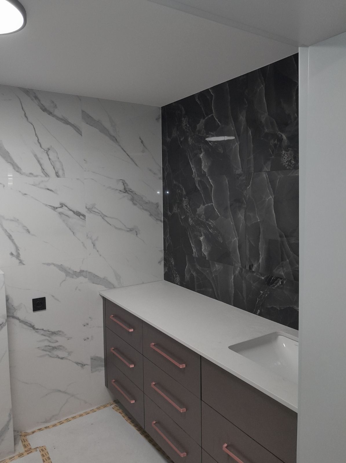 Wykończona łazienka z marmurowymi płytkami w odcieniach bieli i czerni, brązowa szafka z białym blatem i umywalką, oraz miedzianymi uchwytami szuflad.