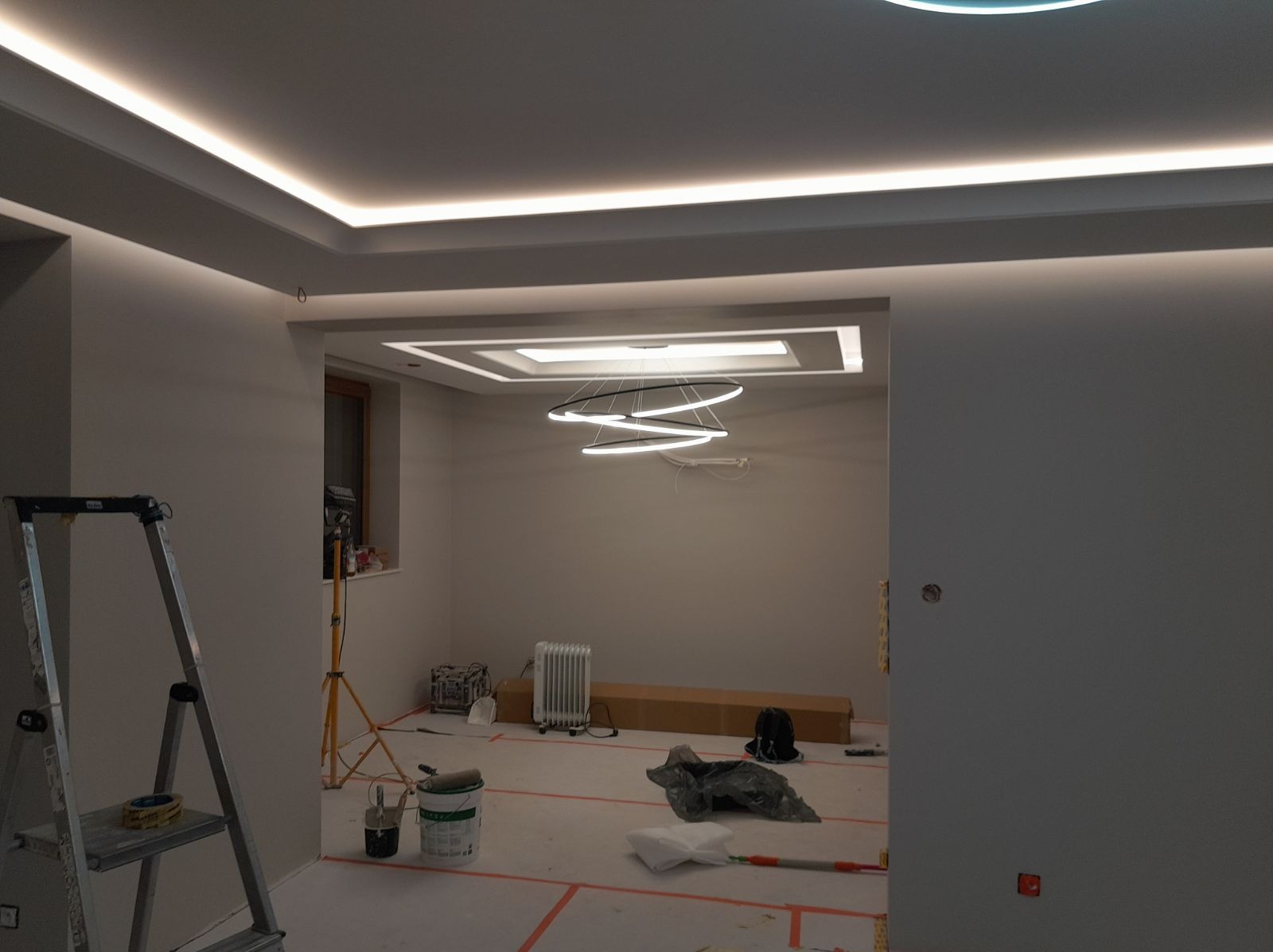 Pokój w trakcie wykańczania: świeżo pomalowane ściany w odcieniach szarości, zabezpieczona podłoga z widocznymi taśmami malarskimi, drabina malarska, wiadro z farbą, grzejnik i nowoczesne...