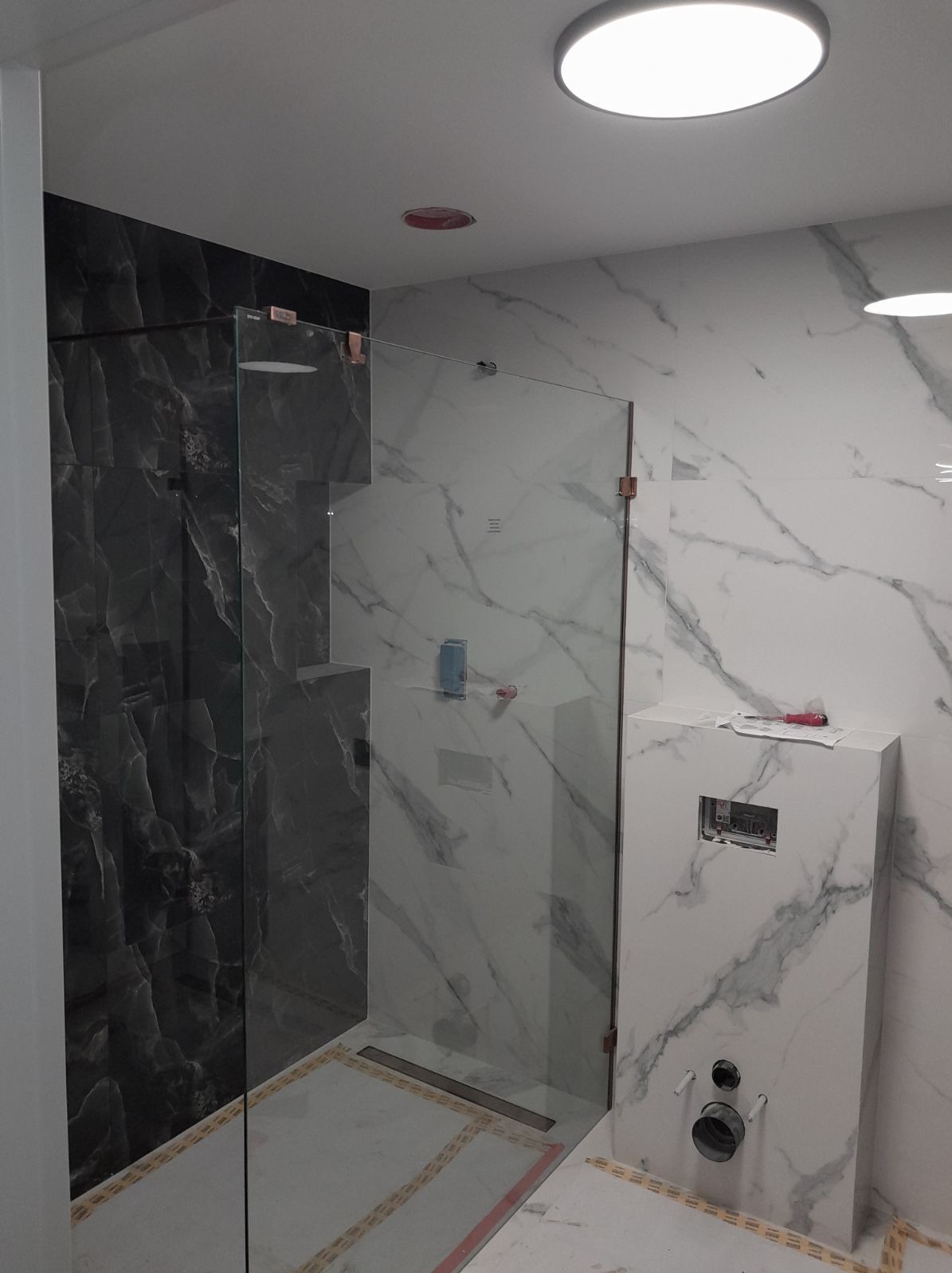 Szklana kabina prysznicowa z marmurowymi płytkami w trakcie montażu, widoczne niedokończone podłączenia hydrauliczne i narzędzia na obudowie stelaża podtynkowego.