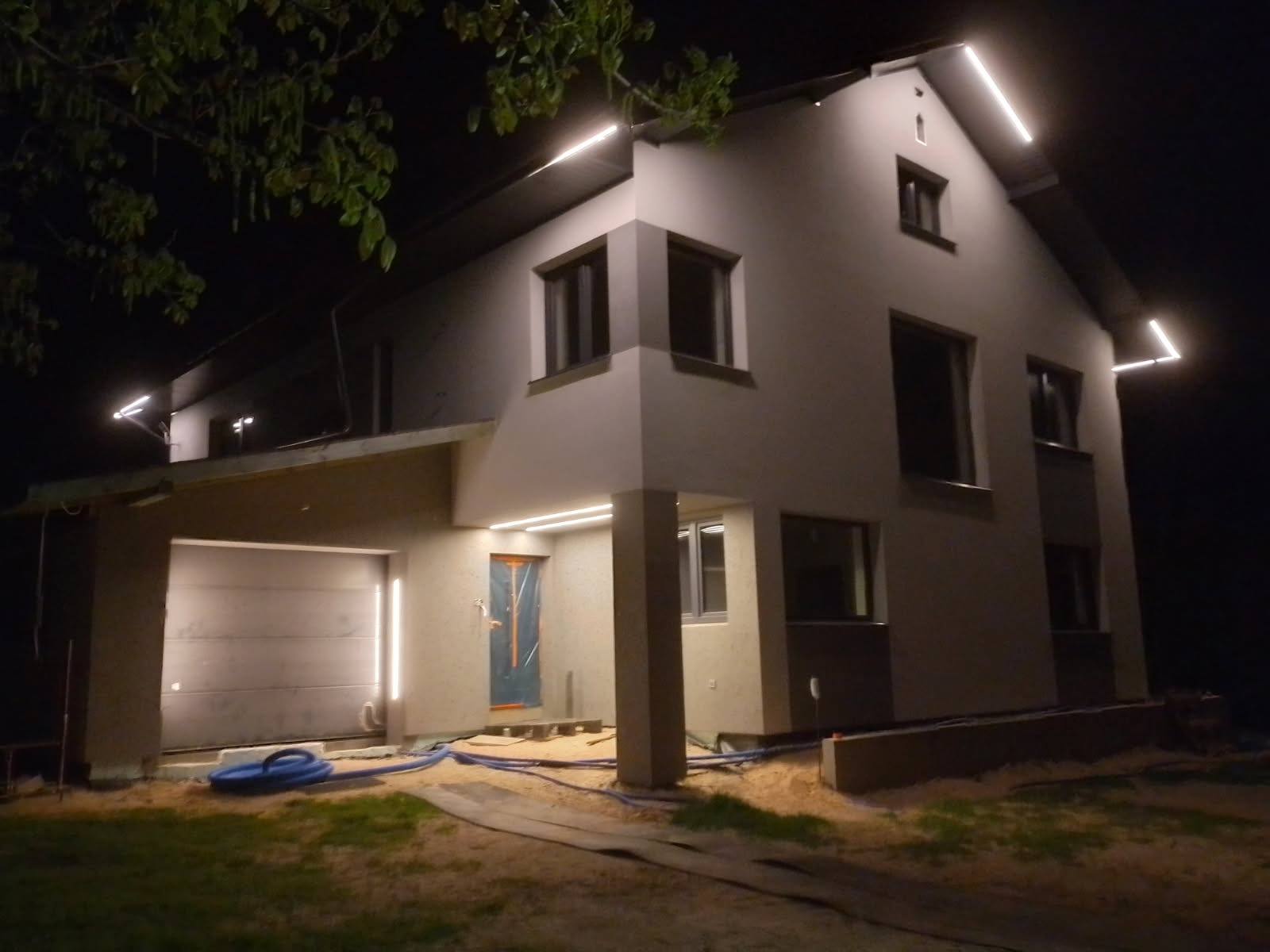 Nowoczesny dom jednorodzinny po renowacji elewacji, nocne ujęcie z oświetleniem LED podkreślającym architekturę. Częściowo zakryte okno folią ochronną.