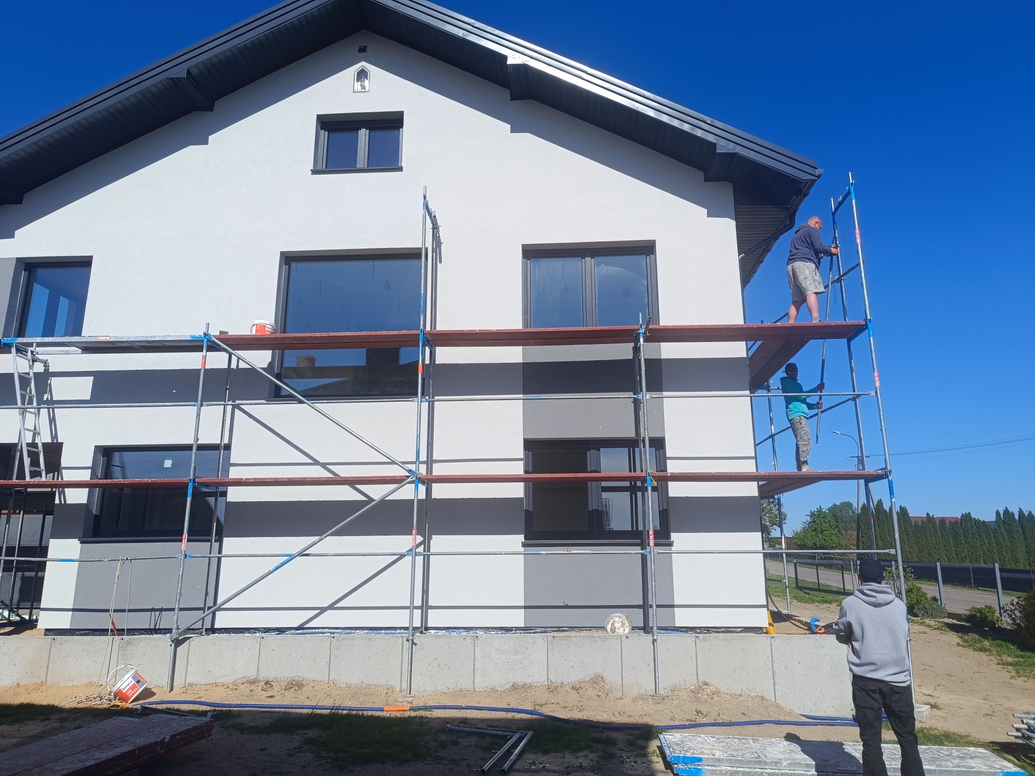 Praca na rusztowaniu przy elewacji domu z szarą stolarką okienną. Widoczny fragment ocieplonej ściany w dwóch odcieniach szarości i pracownicy. Słoneczny dzień.