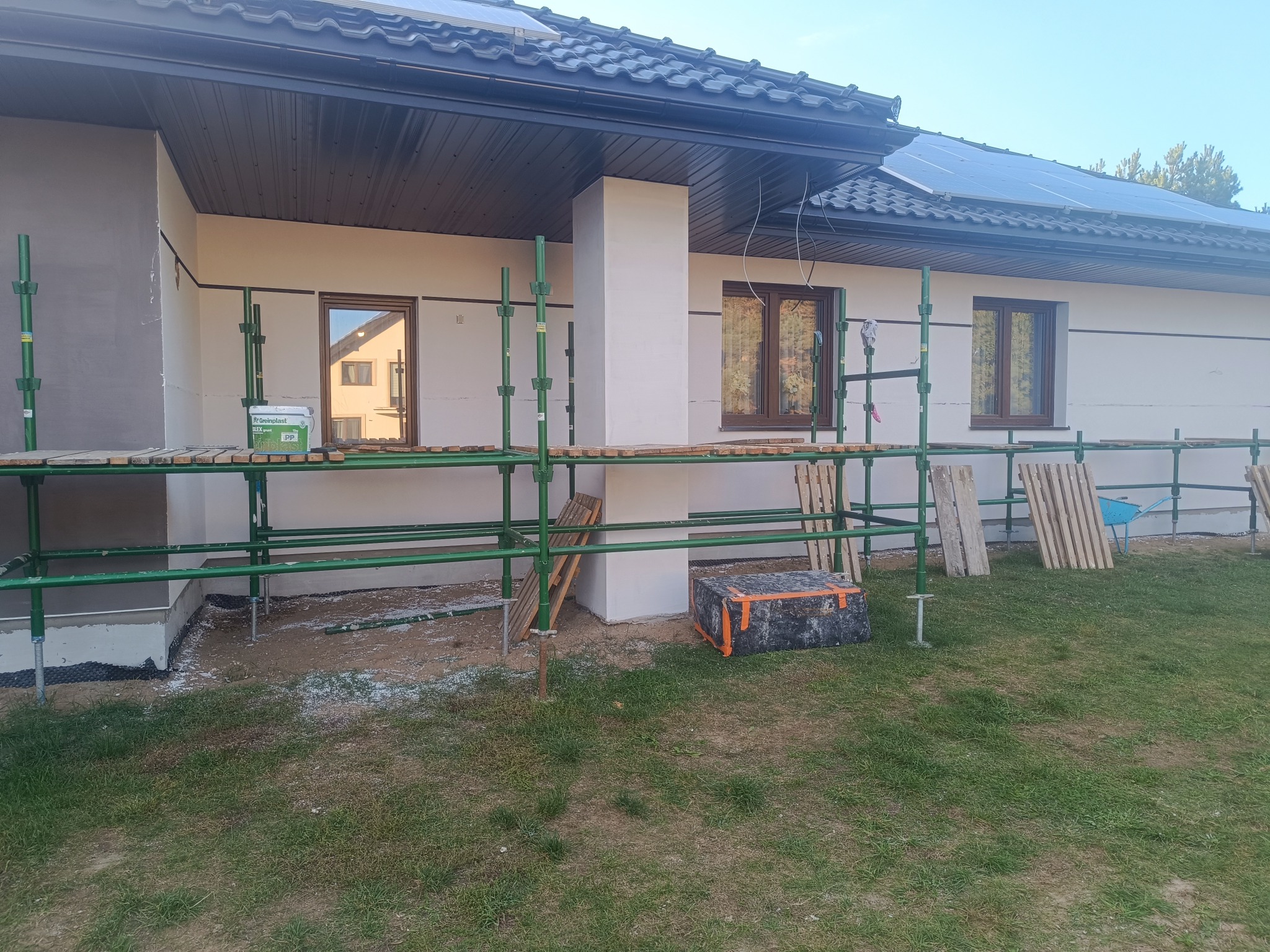 Dom jednorodzinny w trakcie prac elewacyjnych. Widoczne rusztowania, fragment ocieplonej ściany i wiadro z farbą. Na dachu panele solarne.